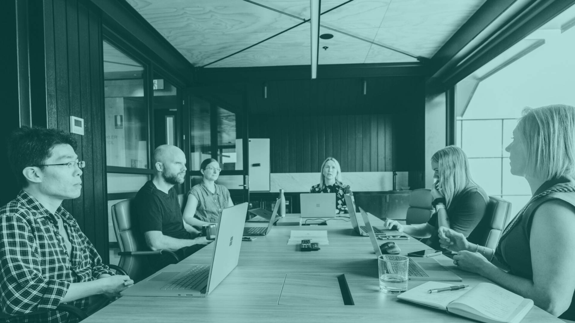 Six people sitting at a boardroom table looking at their laptops